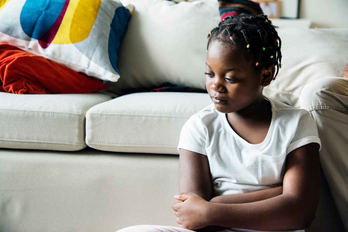A black girl sits quietly at the base of a couch.