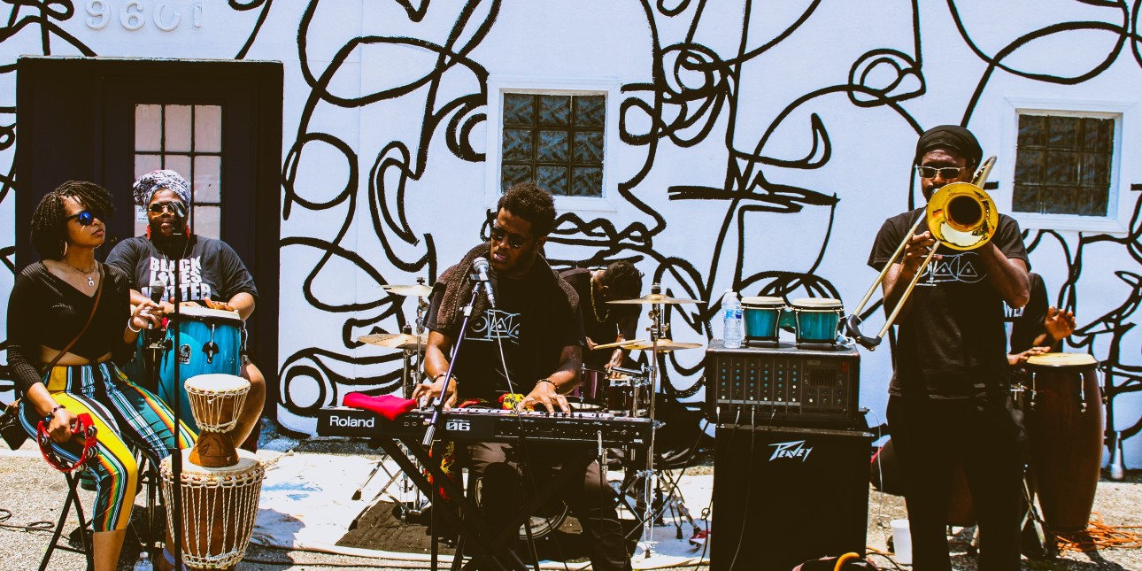 6 Black musicians play music in front of a mural outside