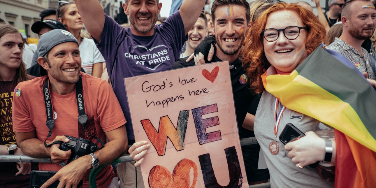 a group of Christians celebrating at Pride with a poster that says "God's love happens here - We 'heart U'"