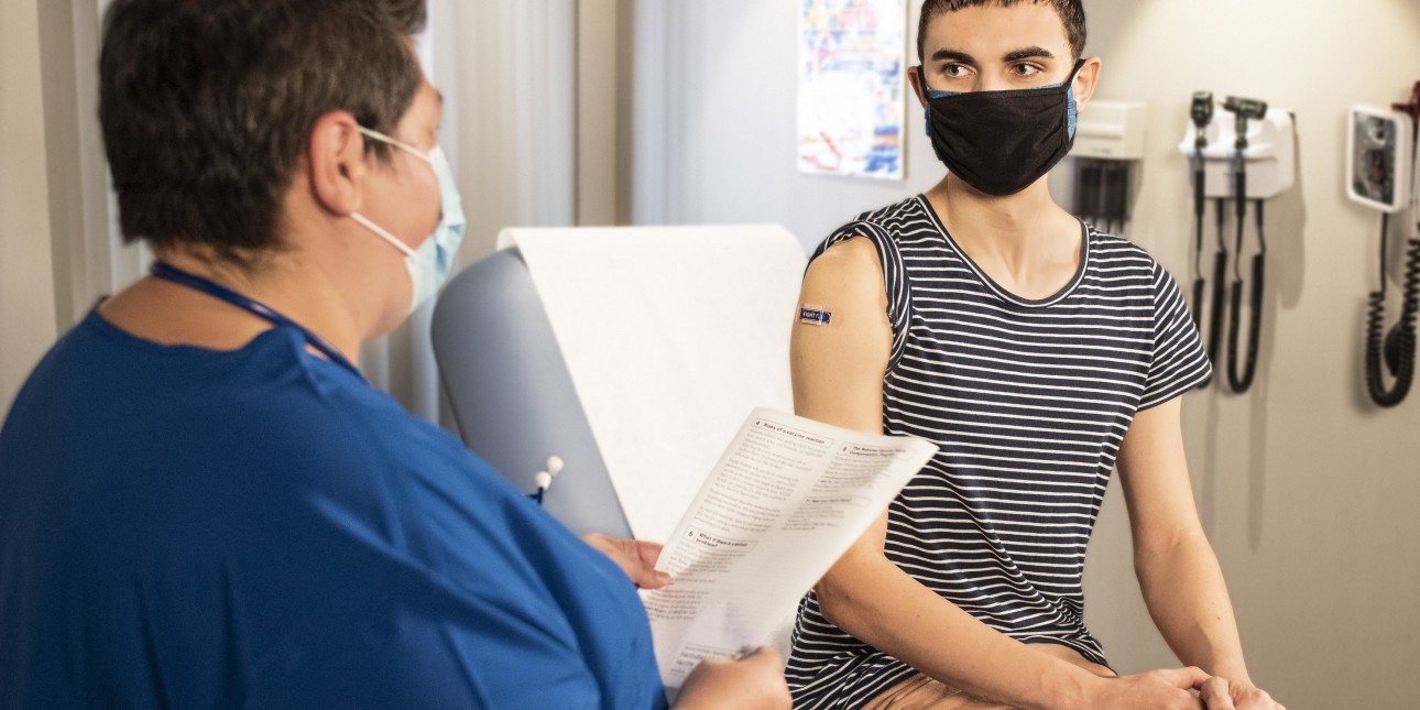 a young person wearing a mask sitting on an exam table talking to medical provider holding a piece of paper