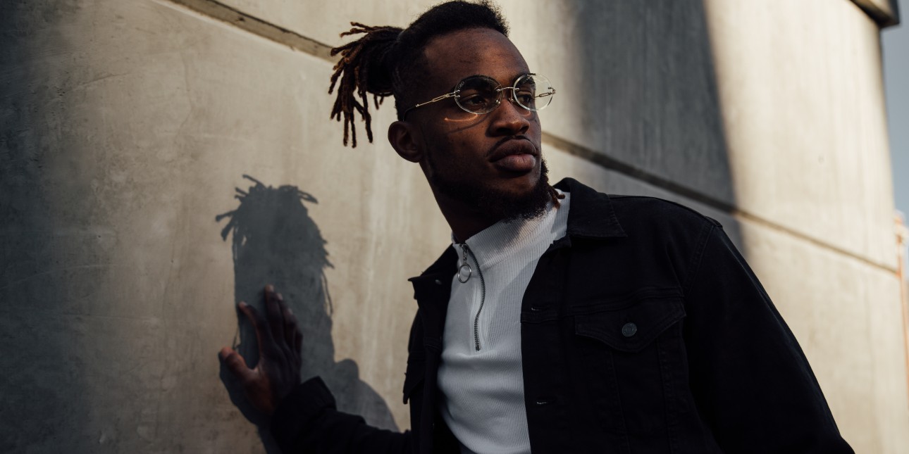 Young adult with dark brown skin, with black hair and goatee, wearing eyeglasses and white half zip shirt with a dark colored jacket standing in front of cement wall with one hand placed on the wall.