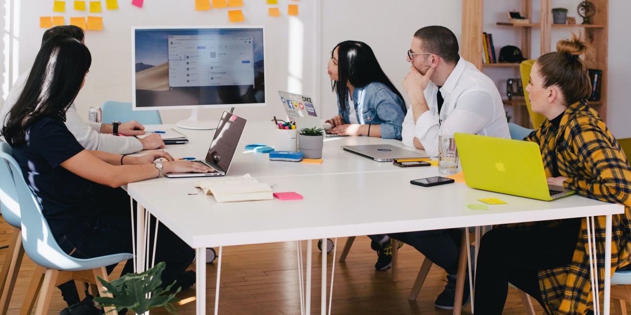 Group of people sitting at table in a collaborative space with post-it notes on a white board and laptops on the table. All looking at a larger computer screen with contemplation. 