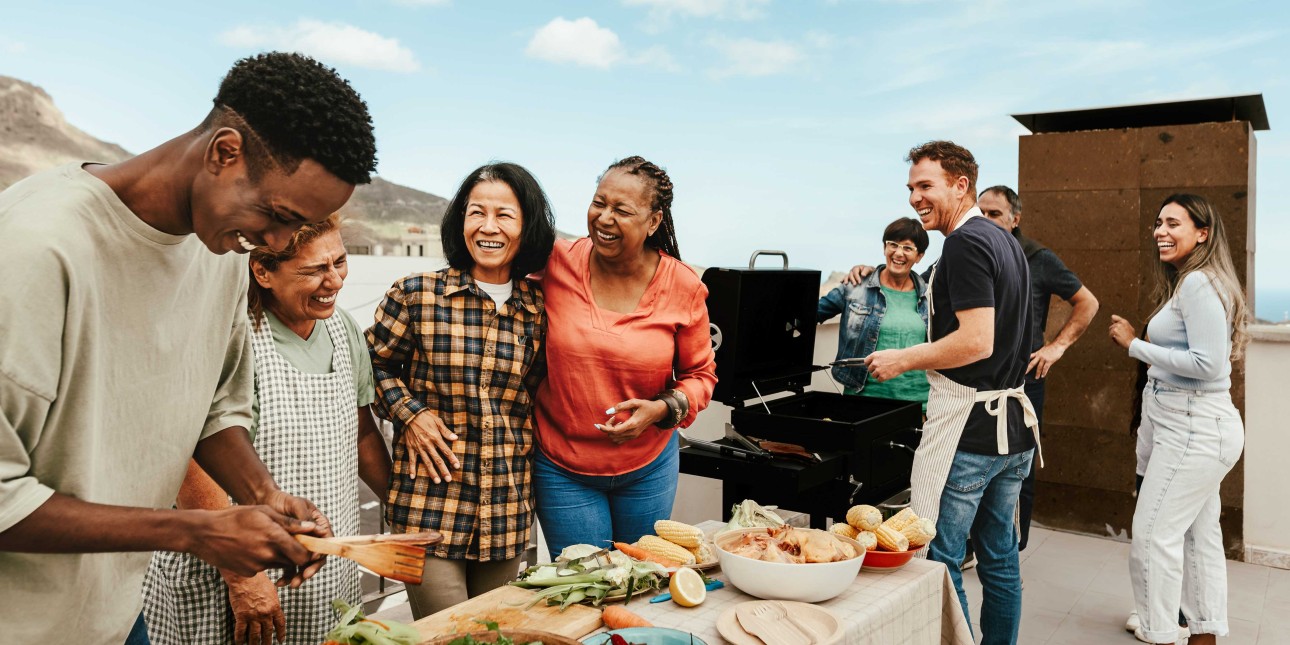 Image of 8 multigenerational people,  smiling and laughing, and with a table of food and a barbecue grill on a rooftop on a sunny day with a bright sky and scattered clouds. 