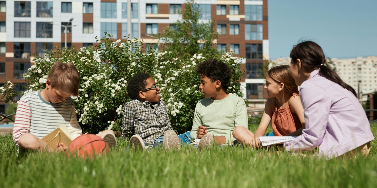 Children sitting together on the grass on a sunny day with trees blooming behind them.