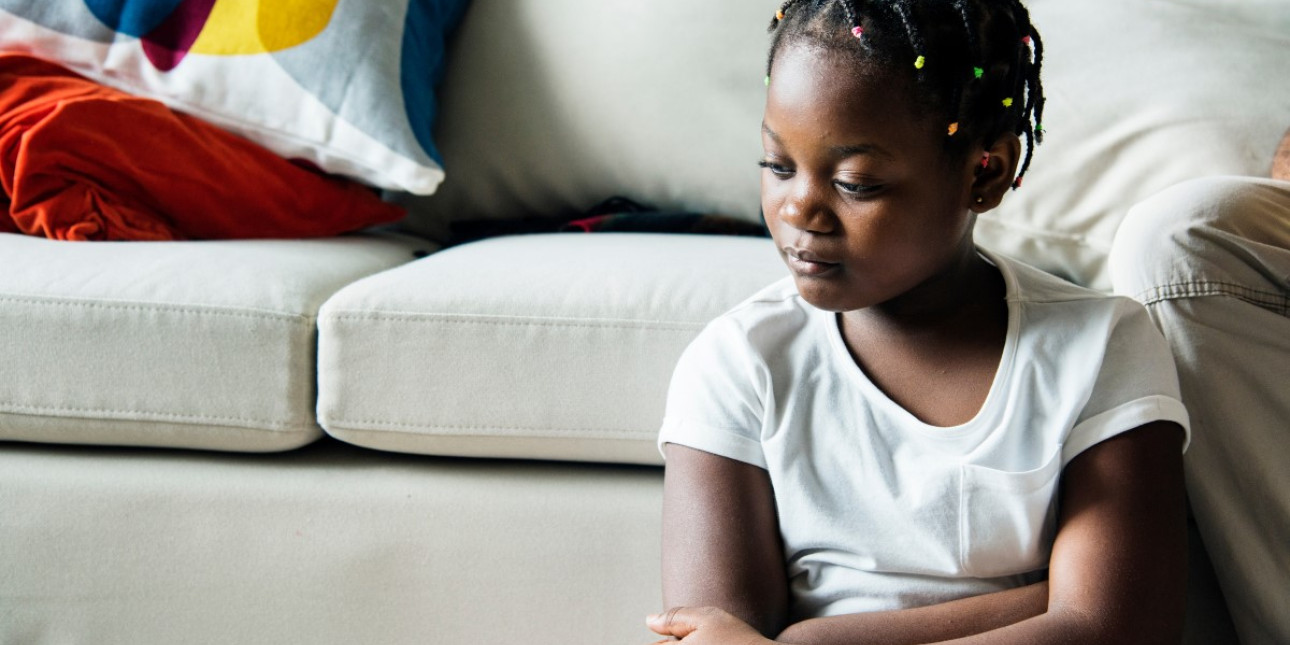 A black girl sits quietly at the base of a couch.