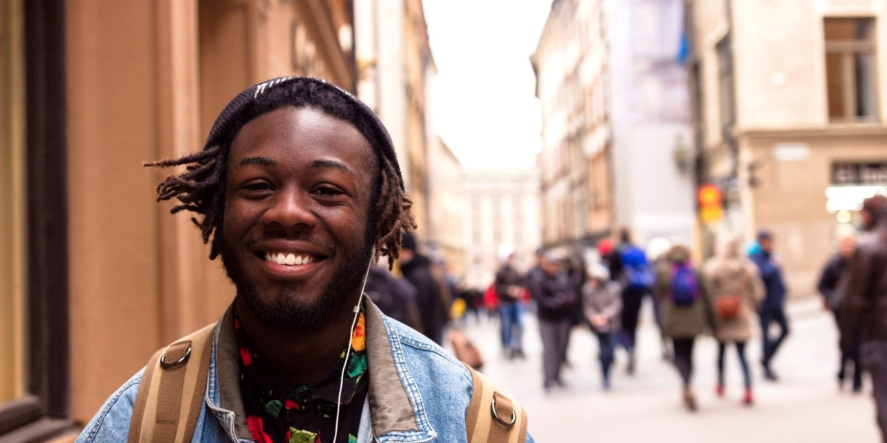 A young smiling person wearing a backpack in focus, a city street with pedestrians in the background