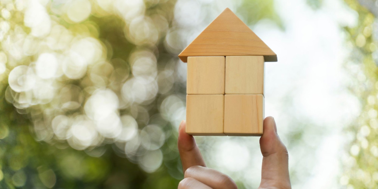 A hand holding up a house made of building blocks against a nature background