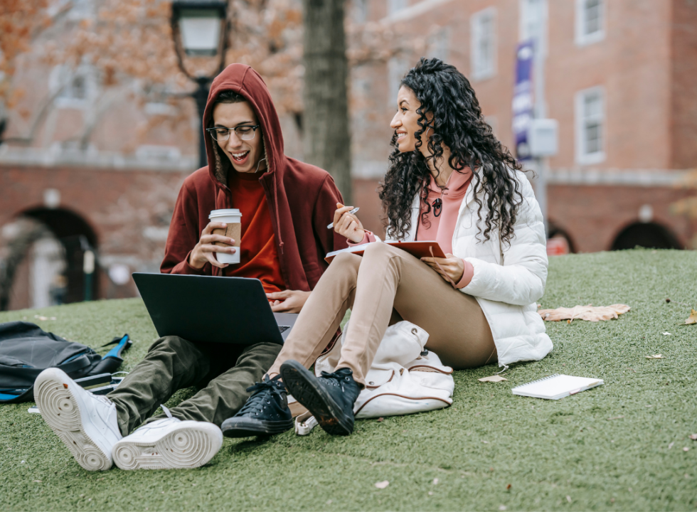 two young people sitting on the ground talking one holding a notebook the other with a laptop and coffee