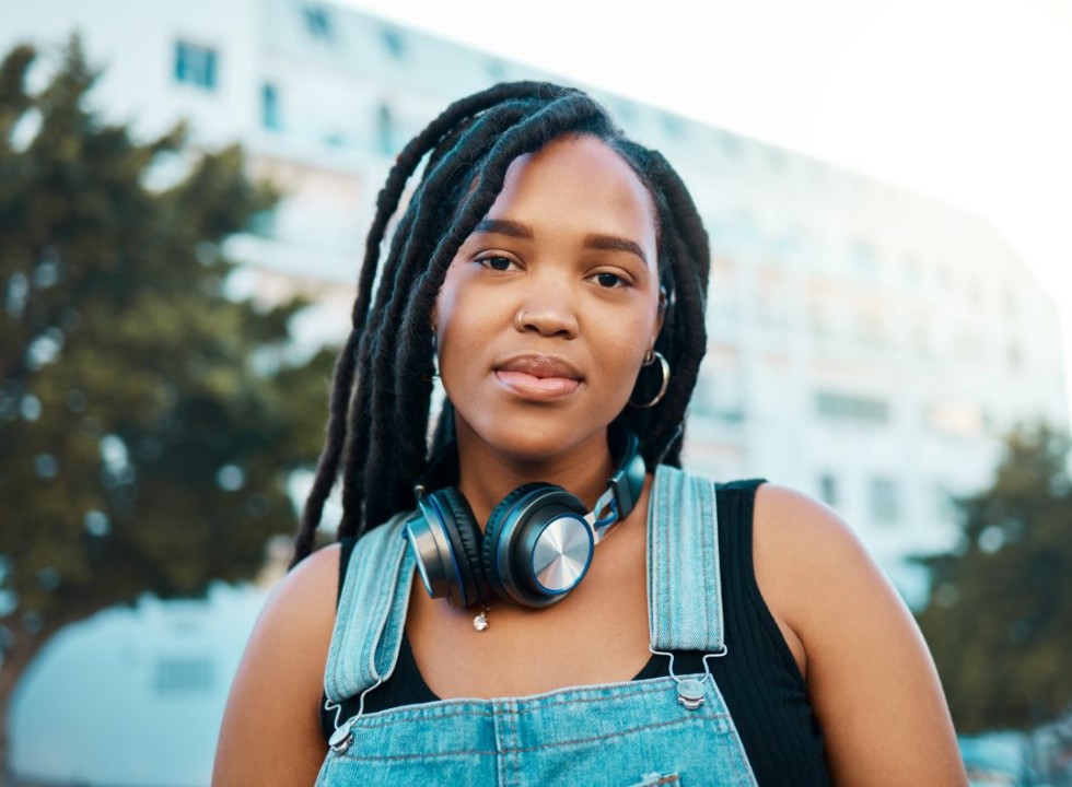 Black woman in a city, wearing headphones around her neck and overalls
