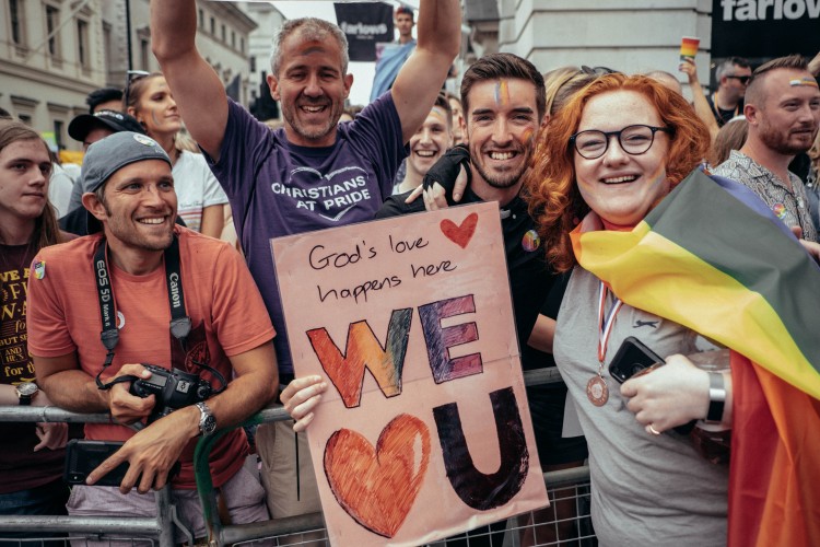 a group of Christians celebrating at Pride with a poster that says "God's love happens here - We 'heart U'"
