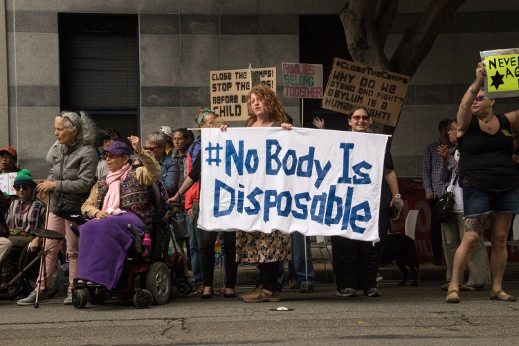 group of protesters - one holding a sign reading "#No Body Is Disposable"