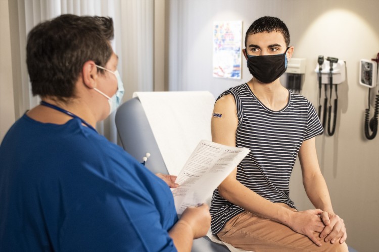 a young person wearing a mask sitting on an exam table talking to medical provider holding a piece of paper