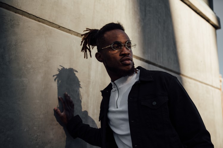 Young adult with dark brown skin, with black hair and goatee, wearing eyeglasses and white half zip shirt with a dark colored jacket standing in front of cement wall with one hand placed on the wall.