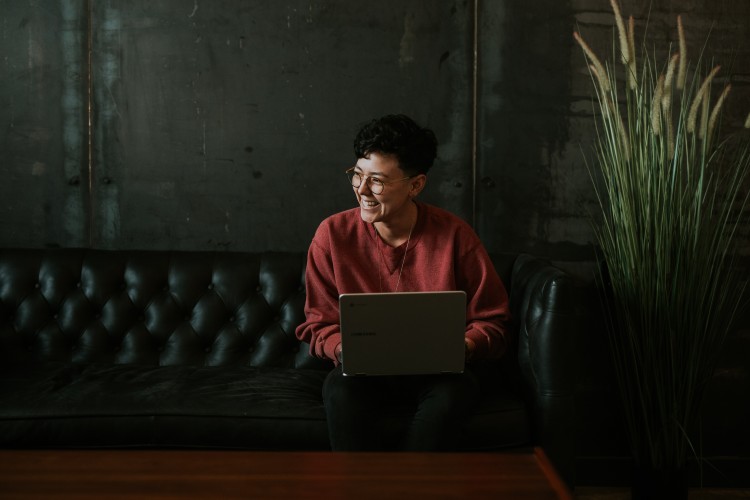 Smiling young adult with tan skin and black hair wearing a red sweater, seated on a couch with a laptop placed on their lap.