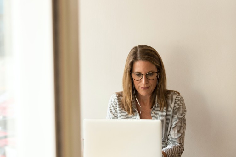 Image of a blonde woman in a blazer seated at a laptop computer in which she is looking at. In front of her on the table is a newspaper.