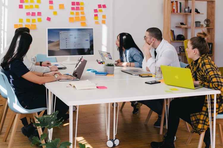 Group of people sitting at table in a collaborative space with post-it notes on a white board and laptops on the table. All looking at a larger computer screen with contemplation.