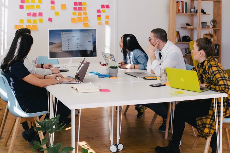 Group of people sitting at table in a collaborative space with post-it notes on a white board and laptops on the table. All looking at a larger computer screen with contemplation. 