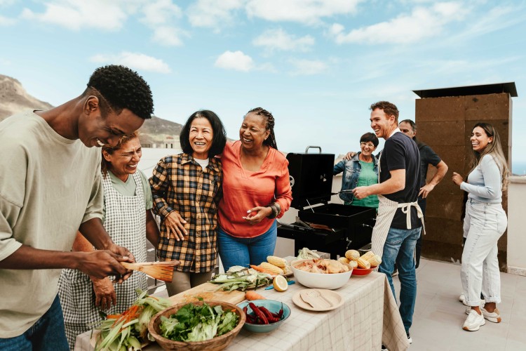 Image of 8 multigenerational people,  smiling and laughing, and with a table of food and a barbecue grill on a rooftop on a sunny day with a bright sky and scattered clouds. 